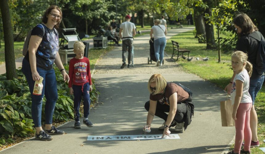 Grupa ludzi jest w parku. Jedna kobieta maluje sprayem na ziemi, podczas gdy inna uśmiecha się do kamery. Obserwuje dziecko w czerwonej koszulce i drugie dziecko w różowej koszuli. Inni ludzie idą w tle obsadzoną zielenią ścieżką.