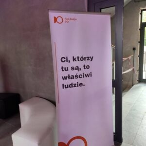 Stojący baner wewnątrz nowoczesnego budynku widnieje z napisem „Ci, którzy tu są, to właściwi ludzie”. u góry posiada logo i nazwę Fundacji DO. Tło jest szare, w pobliżu znajdują się minimalistyczne meble.