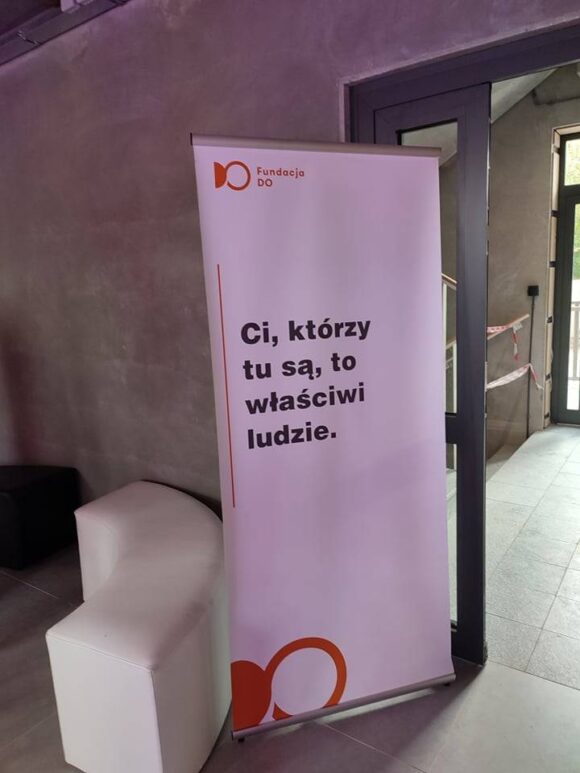 Stojący baner wewnątrz nowoczesnego budynku widnieje z napisem „Ci, którzy tu są, to właściwi ludzie”. u góry posiada logo i nazwę Fundacji DO. Tło jest szare, w pobliżu znajdują się minimalistyczne meble.