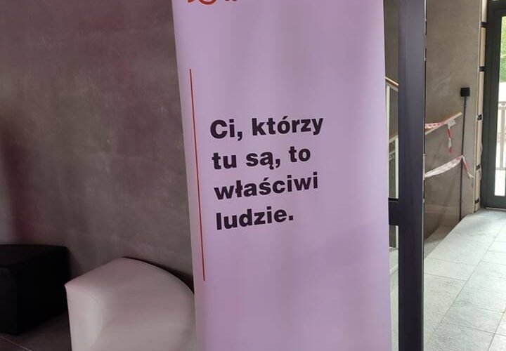 Stojący baner wewnątrz nowoczesnego budynku widnieje z napisem „Ci, którzy tu są, to właściwi ludzie”. u góry posiada logo i nazwę Fundacji DO. Tło jest szare, w pobliżu znajdują się minimalistyczne meble.